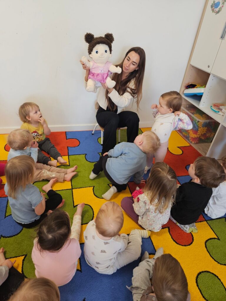 Dzieci siedzą na dywanie wokół pani, która trzyma w dłoniach pacynkę-lalkę.